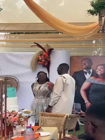 A man in a tan suit is feeding a woman a piece of cake. The woman is wearing a silver dress with beaded accents. Behind them is a sign with the name "Emanuelle" and a picture of them in formal wear. To their left, there is a tiered cake with a blue frosting layer on top and a floral arrangement. They are both smiling. A hand holding a smartphone is visible in the foreground. The scene is decorated with orange and brown accents under a tent.