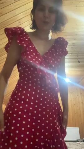 A woman with short brown hair wearing a red and white polka dot dress stands in a room with wood-paneled walls and ceiling. She is looking at the camera and touching her hair.