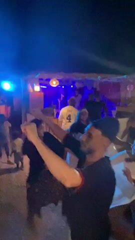 A group of people are dancing at night. The scene is lit by stage lights, with blues and purples dominating. Some people are wearing white clothing, while others are in darker attire. The ground appears to be a dusty or sandy surface. The overall atmosphere is lively and energetic, with a sense of movement and celebration.