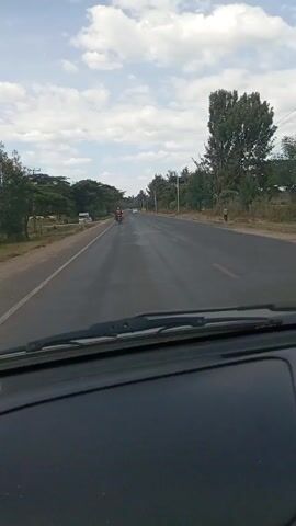 The video shows a car driving on a paved road in a rural area. The sky is blue with scattered white clouds. Trees line both sides of the road. In the distance, a motorbike and a white car are visible. A person is walking on the side of the road. There is a parked white truck on the left side of the road. The video is shot from inside the car, showing the dashboard and windshield.