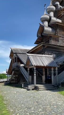 A person walks up wooden stairs toward a large, multi-domed wooden church under a blue sky. The church is ornately decorated with wooden carvings and has a very traditional Russian architectural style. The stairs and wooden structures are weathered and show signs of age. The ground in front of the church is paved with cobblestones. The overall scene depicts a historic wooden church complex on a sunny day.
