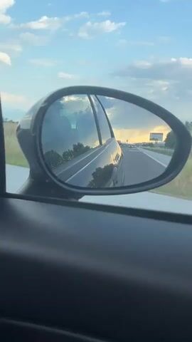 The video shows a view from the side mirror of a car driving on a highway. The mirror reflects the road behind the car, with other vehicles visible in the distance. The sky is cloudy with patches of sunlight breaking through, creating a dramatic effect. Trees and other vegetation are visible along the side of the road. The overall impression is of a scenic drive, possibly at sunset or sunrise, with a sense of movement and the open road.