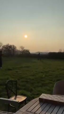 Two people, likely young, play soccer in a large grassy field at dusk or dawn. The sun is low in the sky. One person wears a number 10 jersey. There is a table and chairs in the foreground. Beyond the field is a line of trees and a distant hill or ridge.