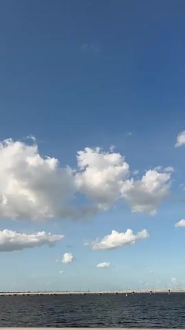 The video shows a partly cloudy sky with a long bridge or landmass in the distance. The sky is a clear, bright blue at the top, gradually fading into a lighter blue where the clouds are scattered.  The clouds are white and fluffy, with some areas of gray shading indicating depth. The bridge or landmass appears as a dark, thin line horizontally across the lower portion of the frame, suggesting a view from a low angle, possibly from a boat.  The water is dark and calm. There is a distinct sound of a horn blowing that is noticeable.