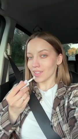 A light-skinned woman with blonde hair and green eyes is applying lip gloss while seated in the driver's seat of a car. She's wearing a white t-shirt, a brown and white plaid shirt, and jewelry. The car seat is black leather, and the woman is wearing a seatbelt. The background shows a glimpse of trees and sky through the car window.