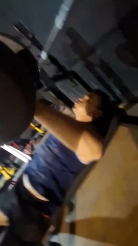 A person is bench pressing a barbell in what appears to be a home gym. They are lying on a bench, lifting the weighted barbell up and down. The room is dimly lit, and workout equipment can be seen in the background. The individual is wearing a blue tank top and black shorts. The camera angle is from below, looking up at the person lifting the barbell.