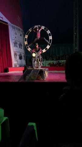 A circus act takes place on a red-carpeted stage under a blue-lit circus tent.  A woman, clad in a black mask and tight black and red clothing, suspends herself inside a large, circular metal prop resembling a clock face. Below her, a man in goggles and a Victorian-esque outfit sits on a motorcycle. The motorcycle is positioned on a black, box-shaped platform with ramps leading up to it.  In the background, an audience is seated and observes the act. To the left of the stage is a backdrop resembling a turreted building.