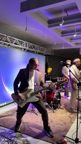 This media file shows a band performing on a stage. The subjects are four men. One man in the foreground is playing a white bass guitar and wearing a brown suit with purple suspenders. To his right, another man in a light-colored suit is playing a guitar. Behind them, a drummer is seated at a red drum kit. Further back, a fourth man, also in a light-colored suit, is playing a keyboard. The setting appears to be an indoor venue with a large LED screen displaying abstract colorful graphics behind the performers. Stage lighting, predominantly purple and blue, illuminates the scene. The floor is covered with a textured rug. The overall atmosphere suggests a live musical performance. The timestamp indicates the video begins at 00:00. The location context provided is Каджи-Сай 2, Kyrgyzstan. No text is visible in the media file itself.