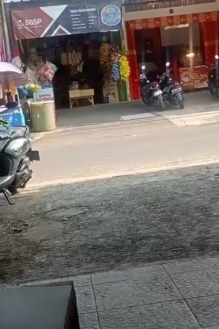 The video shows a street scene in front of two shops. The shop on the left has a sign that says "GGSP TOKO BARANG." It appears to be a general store selling various items, including what look like packaged goods and possibly food items hanging from the ceiling. There is a green trash bin and a motorcycle parked in front of this shop. The shop on the right has a red awning and appears to be a food stall, possibly selling fried chicken given the signage. Several motorcycles are parked in front of this shop as well. The street is paved and there are pedestrians visible in the distance. The sun is shining, casting shadows on the ground.