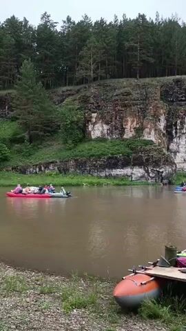 A wide, muddy river flows past a steep, rocky cliff face covered in green vegetation and trees. Several inflatable rafts with people are on the water. In the foreground, an orange and grey inflatable raft is beached on the grassy bank, loaded with bags and paddles. Further out in the river, a pink raft is filled with people, and other smaller rafts with individuals or small groups are visible. The sky is overcast.