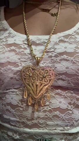 A person is featured in a close-up, showcasing multiple gold necklaces over a white, semi-sheer lace top. The most prominent necklace features a large, ornate golden heart-shaped pendant adorned with intricate filigree flowers and small tear-drop danglers. Several other delicate gold chains are layered around the neck, one clearly displaying the numbers "333" and another with the word "ANGEL" as pendants. The camera zooms out slightly to reveal a glimpse of a green, patterned fabric, possibly a scarf or skirt, below the waist. The setting appears to be indoors, with ambient lighting, and music is audible in the background.