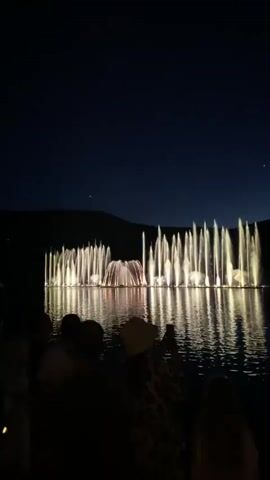 A nighttime outdoor scene features a synchronized water fountain show with illuminated water jets creating dramatic vertical plumes against a dark sky. A body of water reflects the lights of the fountains. In the foreground, silhouetted figures of people are visible from behind, watching the spectacle. One person in the middle foreground is wearing a hat and holding up a smartphone to record or photograph the event. The overall environment appears to be a lake or large pond with hills or mountains visible in the distant background.