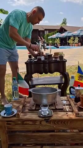 A man in a teal t-shirt and grey shorts prepares coffee using a manual espresso maker on a rustic wooden table outdoors. He is standing on grass in front of a swimming pool and a building with an outdoor seating area. The coffee-making setup includes a large metal pot, coffee beans scattered on a wooden surface, and two small cups. Flags of Italy and Moldova are visible, as is a bottle of water and a moka pot. The man is actively engaged in operating the espresso machine, suggesting a demonstration or a personal preparation of coffee. The overall scene conveys a relaxed outdoor atmosphere with a focus on the coffee-making process.