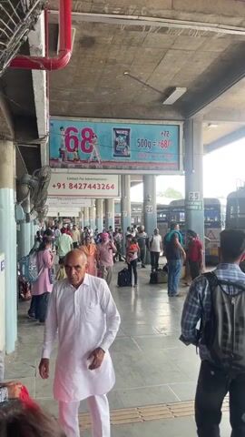 The video shows a busy bus terminal, likely in India, filled with people walking around. The camera pans right, revealing several buses parked in designated bays labeled with numbers, such as 25 and 24. The buses have text written in a language other than English, potentially Punjabi, along with advertisements and illustrations. There are advertisements above on the building. Passengers appear to be waiting for their respective buses.