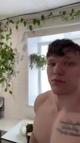 A shirtless young man with a tattoo on his left arm flexes his bicep, looking directly at the camera. He is standing indoors in front of a white countertop with glass jars on it. Behind him is a window with a view of a snowy exterior and other buildings. A leafy green plant hangs from the ceiling in the top left corner of the frame.