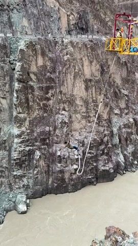 A person is bungee jumping off a cliff into a brown river. The bungee jumper is suspended by a rope. There is a platform attached to the cliff face from which the jump is made. The cliff is rocky and steep, and the river is wide and appears to be fast-flowing. The weather is overcast.