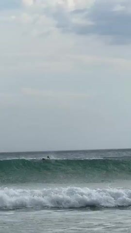 The video shows ocean waves breaking on a shore on a cloudy day.  The waves are green-blue and foamy as they approach the shore.  There is a distant figure in the water, possibly a surfer. A bird flies in the distance, briefly visible in the sky. The sky is mostly overcast with grey clouds.