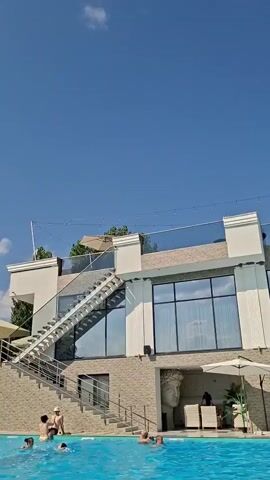 A wide shot of a poolside bar with people swimming in the foreground and sitting at tables under umbrellas. A large sculpture of a head is visible on the side of the building. The sky is blue with some clouds. The video pans slightly to the right.