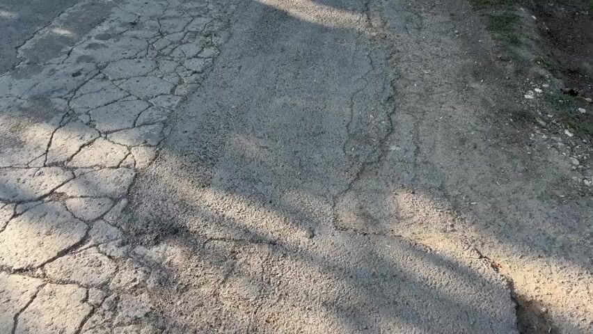 The video shows a cracked asphalt road in a village. The road is lined with fences and trees. The sun is shining brightly and casting shadows on the road. A car is parked in the distance.