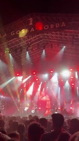 A low-angle shot captures a live band performing on a large stage during a concert. The stage is brightly lit with red and white spotlights that cut through the smoke-filled air. The band members are silhouetted against the bright lights, with some identifiable playing instruments like guitars and drums.  A large structure with a number one and the words "LA GOSSA SORDA" is clearly visible on the stage. In front of the stage, a crowd of people is visible, facing the band with their backs to the camera, indicating they are watching the performance. The overall atmosphere suggests a lively and energetic concert experience.