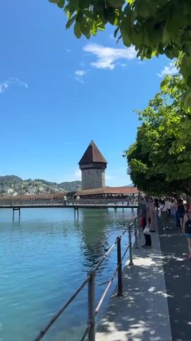 This video shows a picturesque waterfront scene with a prominent octagonal tower and a covered wooden bridge spanning across a body of water. The water is a vibrant turquoise color, reflecting the clear blue sky above. Along the right bank, a paved walkway is lined with a metal railing and shaded by lush green trees. Several people are visible strolling along the walkway. In the background, a charming European town with distinctive architecture, including spires and red-tiled roofs, is nestled against rolling green hills. The sun is shining brightly, indicating a pleasant day. The camera pans slowly to the left, capturing the expanse of the water and the town.