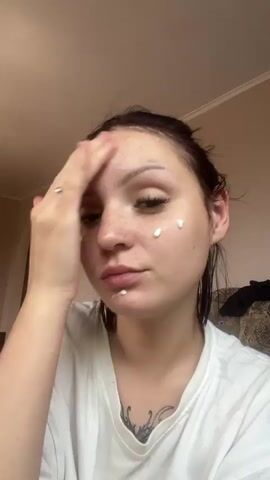 A young woman with her hair pulled back applies cream to her face. She is wearing a white t-shirt. Several dots of cream are visible on her cheeks and forehead, and she is rubbing the cream into her skin with her right hand. She has her eyes closed. The background appears to be an indoor setting with a wall and a doorway visible.