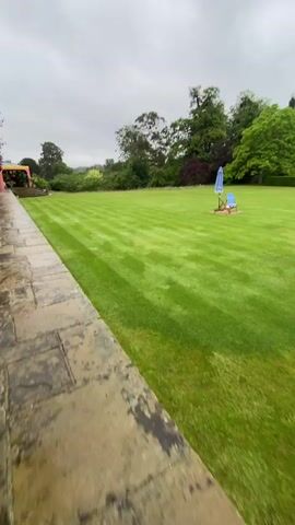 The video pans across a manicured green lawn with striped patterns from recent mowing. In the foreground to the right, two blue and white striped deck chairs are set up on a brown mat, with a blue umbrella. Further out on the lawn are a few more deck chairs and umbrellas. In the background, a large, stone house with multiple gables and chimneys is visible. There are people walking in front of the house. The sky is overcast.