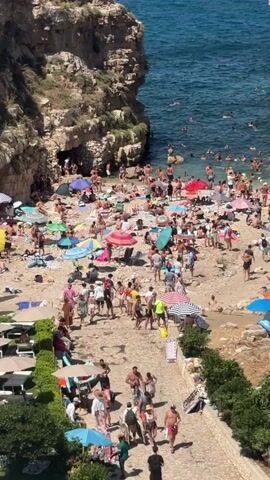 This aerial view captures a densely crowded beach on a sunny day, featuring a large number of people engaged in various beach activities. The beach is situated in a cove, with rocky cliffs on either side and a town built on the cliff edge on the right. The water is a vibrant blue-green, with many individuals swimming and floating. The beach itself is a mix of sand and pebbles, covered with numerous beach umbrellas in various colors, beach chairs, towels, and people lounging. People are scattered across the beach, some walking, some sitting, and some interacting with each other. A pathway runs along the bottom of the frame, bordered by greenery, with more people walking along it. The overall scene depicts a popular and busy tourist destination during peak season.