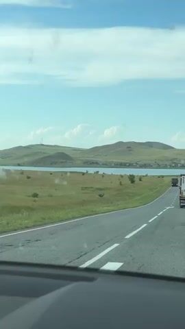 The video begins with a view of a winding road through a rural landscape. Rolling green hills with some scattered trees are visible in the background, with a body of water in the middle ground. The sky is blue with some scattered clouds. As the video progresses, the camera pans slightly to the right, revealing a few trucks traveling on the same road in the same direction. The video then shows a reflection in the side mirror of a car. In the reflection, a person is visible holding a smartphone and taking a selfie. The background in the mirror reflection is similar to the landscape outside the car, with green hills and a blue sky. The video ends with the car continuing down the road.
