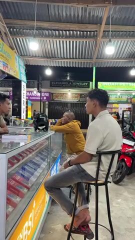 This media file captures a candid scene at what appears to be a small electronics and service shop in Bireuen Meunasahreuleut, Indonesia. Three men are the central subjects. On the left, a man in a light brown t-shirt is seated behind a counter, appearing to be engaged in some activity on the counter, possibly paperwork or arranging items. In the center, another man, wearing a mustard yellow hoodie and dark pants, is leaning on the counter, looking towards the man on the right. He seems relaxed and engaged in conversation. On the right, a man in a light-colored polo shirt and gray jeans is seated on a tall stool, facing away from the camera and towards the other two men, also appearing to be part of the conversation. A black helmet rests on the counter between the man in the yellow hoodie and the man behind the counter.

The setting is an indoor/outdoor space, indicated by the corrugated metal roof supported by wooden beams overhead. Fluorescent lights illuminate the area, suggesting it might be evening or nighttime, or a dimly lit indoor space. The counter itself is a glass display case filled with numerous boxed items, likely mobile phones, given the branding visible on the counter itself and the signage in the background.

Visible text includes "BSI AGEN JUANG CELL 2" on a yellow sign, and "BSI" is prominently featured in orange and white on the counter's front. Other signs in the background indicate businesses like "ELF IJUMELA JEWELRY" and "INDAH PETSHOP," suggesting a commercial area with multiple small shops. The presence of a red motorcycle parked on the right side of the frame further reinforces the Indonesian setting and the likely mode of transportation in the area.

The overall atmosphere seems casual and conversational, with the men appearing comfortable and relaxed in their interaction. The lighting, the attire of the individuals, and the type of businesses visible point to a typical urban or semi-urban commercial setting in Indonesia, likely after business hours.