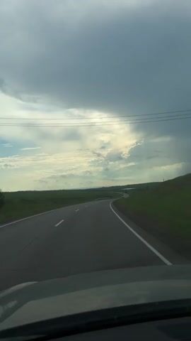 The video shows a view from inside a vehicle driving on a highway. The highway curves to the right and is bordered by green fields on both sides. The sky is overcast with dark clouds, but there is a break in the clouds where sunlight is shining through. There are a few other cars visible on the road. The overall impression is of a slightly gloomy but open landscape.