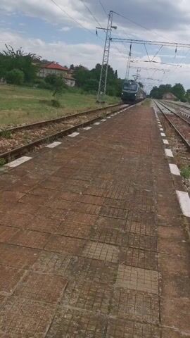 The video shows a train traveling along a train track, as seen from a platform. The train has a green and blue locomotive with the words "BDZ" on the side. The platform is made of brown tiles with white edges. There is grass and a building to the left of the tracks. The sky is cloudy. The camera is stationary and follows the train as it passes by.