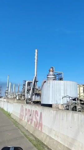 From a moving vehicle on a sunny day, the video shows a petroleum refinery with large tanks, pipes, and smokestacks against a clear blue sky. The foreground features a concrete wall with the "PDV" logo painted on it. No people are visible in the video. The camera pans across the industrial landscape, showcasing various structures within the refinery complex. The overall atmosphere is industrial.