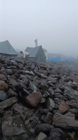 The video shows a rocky landscape with several tents pitched on it, shrouded in heavy fog. The ground is covered in dark gray rocks, many of which are wet.  The tents are in various colors, including blue, green, orange, and yellow. Several indistinct figures can be seen moving around the tents, appearing as silhouettes in the heavy fog. The fog is thick, obscuring much of the background and giving the scene a low-visibility, somewhat desolate appearance.