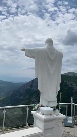 A video captures the back view of a white statue, likely depicting Jesus Christ, standing on a pedestal. The statue has its arms outstretched, overlooking a vast mountainous landscape. Below, deep valleys are visible with a winding river or road snaking through them. The sky is partially cloudy with patches of blue. The statue is positioned on what appears to be a viewing platform, indicated by a modern railing with glass panels in the foreground. Artificial flowers in shades of blue and green adorn the base of the statue, and a small white device sits on the railing to the left. The camera slowly pans slightly to the right, revealing more of the dramatic mountain scenery. The setting is outdoors, likely at a high altitude, providing a panoramic view of the natural environment in Cijevna, Montenegro. The time of day is bright, with daylight illuminating the scene. No discernible text is visible in the video.