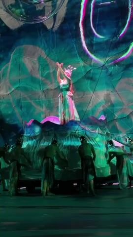 This media file captures a performance scene, likely a theatrical or dance production, set in Nan'an District, China. The foreground features a group of performers dressed in what appear to be ancient or ceremonial attire, with darker, simple tunics and lighter draping fabric. They are positioned around a raised, ornate platform that resembles a stylized water feature or a large bloom, adorned with pink and green accents.

On top of this platform stands a central figure, a performer dressed in an elaborate, flowing gown with iridescent qualities, suggesting a costume of royalty or a mythical being. This central performer wears a detailed headdress and is engaged in graceful, expressive hand and body movements, appearing to be the focal point of the performance. The backdrop is a large, textured screen or curtain illuminated with vibrant projections of swirling, abstract patterns in blues, greens, and purples. These projections evoke a sense of an underwater or ethereal environment, with luminous circular shapes and flowing lines that add to the dreamlike atmosphere.

The lighting is dramatic and theatrical, with spotlights highlighting the performers and the colorful projections creating a magical and immersive ambiance. The overall emotion conveyed is one of artistry, mysticism, and dramatic storytelling, as the performers' actions and the visual design coalesce to create a visually striking tableau. There is no visible text in the media file. The scene appears to be taking place indoors, in a performance venue, given the controlled lighting and stage setup.