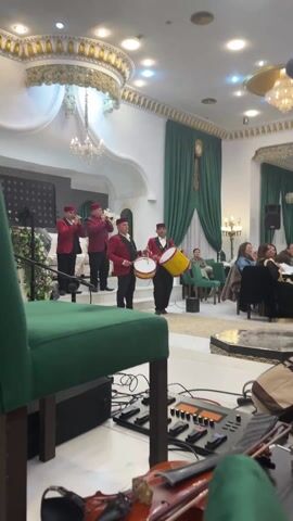 The video shows an indoor scene featuring a musical performance by a group of four men. They are dressed in matching attire, consisting of red jackets with black lapels and traditional dark red, possibly felt, hats resembling a fez. Two of the men are playing wind instruments that appear to be oboe-like or zurnas, holding them vertically and blowing into them. The other two musicians are playing drums; one holds a large yellow drum with a red rim, and the other holds a slightly smaller white or light-brown drum, both suspended from their bodies and played with sticks. They are standing on a raised platform or stage area.

The setting is an elegant room, likely a restaurant, banquet hall, or hotel lounge. The walls are predominantly white with intricate gold detailing along the top edges and on the ornate ceiling. Large chandeliers hang from the ceiling, providing ambient light, supplemented by recessed lighting. Green velvet drapes adorn large windows or wall sections in the background, and there's a tall, old-fashioned clock on the wall between the drapes. Guests are visible seated at tables and in green armchairs in the background, observing the performance.

In the immediate foreground, partially visible, is the back of a green upholstered chair or stool, suggesting the viewer is seated. Below it, on the floor, there's audio equipment, including what looks like a mixer or effects pedal board with several knobs and cables. Next to this equipment, a violin or a similar stringed instrument is resting, partly visible. The overall atmosphere appears to be a formal or semi-formal event with live traditional music entertainment.