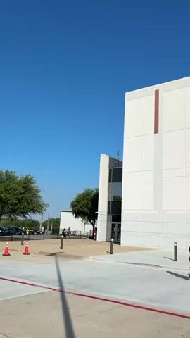 The video shows the exterior of a modern building with a cross on top and the words "The Potter's House" written on the facade. The building appears to be a church or religious institution. Several people are seen walking around the entrance and in the surrounding plaza. The sky is clear and blue, suggesting a sunny day. The architecture of the building is contemporary, featuring large windows and geometric shapes. The overall scene conveys a sense of community and activity.