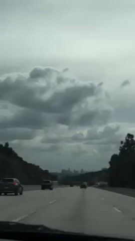 The video shows a view from inside a car driving on a highway. The sky is overcast with thick, gray clouds. The highway has multiple lanes of traffic going in the same direction, with several cars visible in front of the camera car. Trees and hills line either side of the highway. In the distance, the skyline of a city can be seen. The dashboard of the car is visible at the bottom of the frame. The overall scene is somewhat gloomy due to the overcast sky.