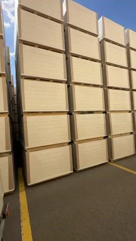 This media file features a large outdoor storage area filled with stacks of pale beige panels. The panels are uniformly sized and neatly stacked on wooden pallets, creating towering walls of material. They appear to be new and are wrapped in plastic or protective film. The scale of the stacks suggests a commercial or industrial setting, possibly a warehouse or distribution yard.

The scene is set outdoors under a bright blue sky with a few wispy white clouds, indicating fair weather and likely daytime. The ground is asphalt, marked with yellow lines, suggesting a parking lot or a designated outdoor storage area within a larger facility. In the distance, a hint of buildings and possibly trees can be seen, providing a backdrop to the industrial scene. There are no people visible in the media file. The overall impression is one of order and large-scale inventory. No text is visible in the media file.