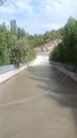 The video shows a canal with water flowing through it. The canal is lined with concrete and has metal railings on either side. Trees and vegetation are visible on the banks of the canal, leading up to a rocky hillside in the background. At the far end of the canal, there is a drop where the water flows over a spillway, creating a waterfall effect. The sky is partly cloudy. There are no people visible in the video.