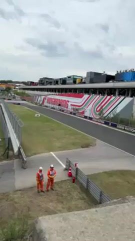 The video shows a Formula 1 race taking place at the Hungaroring. Two people in orange coveralls and white hard hats stand next to a concrete barrier along a section of the track where race cars are speeding past. The track is bordered by a green field on one side and a gray guardrail and fence on the other. In the background, there is a large sign that says "Hungaroring" in large white letters, along with a colorful design featuring red, white, and green stripes. A building with multiple levels and an area for spectators is visible in the background, along with a cloudy sky. The video captures the high-speed action of the race, with the roar of the cars audible.