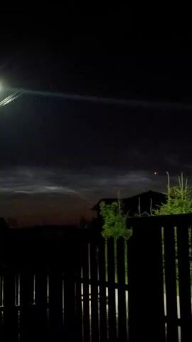 A vertical video captures a dimly lit outdoor scene at night. The foreground is dominated by a dark, wooden fence with vertical planks. Beyond the fence, silhouetted houses and evergreen trees are visible. The sky is dark with some wispy clouds illuminated by a distant light source, possibly the moon or streetlights, creating a luminous effect. A prominent streetlamp on a wooden pole is visible on the left side of the frame, emitting a bright light that casts a glow and creates lens flare. Another smaller light source is visible in the distance on the left. The overall atmosphere is calm and serene.