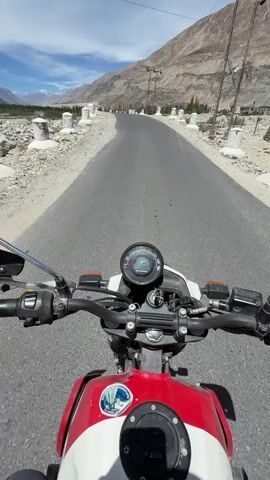 The video shows a first-person perspective of someone riding a motorcycle on a paved road through a mountainous landscape. The rider's hands are visible on the handlebars. The motorcycle is white and red with stickers on the fuel tank. The road curves ahead, leading into a valley with mountains on either side. The sky is blue with some clouds. There are white posts lining the side of the road. The overall impression is one of a scenic motorcycle journey.
