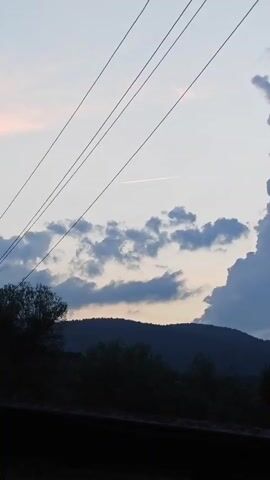 This media file shows a scenic outdoor view, likely captured in Altayskoye, Russia. The image is dominated by a twilight sky, with a mix of soft pinkish hues near the horizon and a pale blue higher up. A few scattered clouds, tinged with grey and blue, drift across the sky, suggesting a temperate weather condition. In the foreground, silhouetted against the sky, are the dark outlines of trees and a sloping hillside, indicating a natural, possibly rural or mountainous, setting.

Stretching diagonally across the upper portion of the frame are several dark lines, which appear to be power lines. Their presence suggests human infrastructure within the otherwise natural landscape. High in the sky, a thin, bright white contrail from an airplane stretches across the frame, marking the path of an aircraft. This contrail, illuminated by the setting sun, adds a dynamic element to the otherwise still scene.

The time of day is clearly evening, given the soft, diffused light and the colors in the sky. The overall mood conveyed is one of tranquility and vastness, with the natural elements of the sky and landscape taking center stage, punctuated by the man-made power lines and the fleeting presence of the airplane. There is no visible text in the media file.