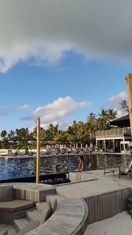 The video captures a tropical resort scene. In the foreground, a curved wooden deck features a built-in lounge area with plush cushions and a small table. Beyond this, a large swimming pool with dark water is visible, with several people swimming and relaxing. Palm trees and lush vegetation line the background, with some buildings and umbrellas also present. The sky is blue with scattered white clouds, suggesting a bright, sunny day. In the middle ground, a small stage covered in thatched material is set up on the beach. Three individuals are on the stage, appearing to be a musical band, with one playing a keyboard and another a drum. Two large speakers are positioned on either side of the stage. The sun is setting or rising in the background, casting a warm glow over the scene.