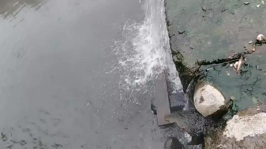The video opens with a close-up shot of murky water flowing over a concrete edge, creating a small waterfall. The water appears somewhat dirty, with debris visible on the bank. As the camera pans down and to the left, the scene expands to reveal a wider view of a slow-moving river or stream. The riverbank is lined with rocks and patches of green vegetation, including grass and trees. The water itself is a dull gray color and shows some ripples from the flow. There are no people or animals visible in the video. The overall impression is a natural, albeit somewhat unkempt, riverside environment.