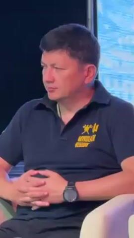 A man wearing a dark polo shirt with a logo on the left breast is seated in a white armchair, looking off to the side. His hands are clasped in his lap, and he is wearing a black watch on his left wrist. Behind him is a blurry screen.