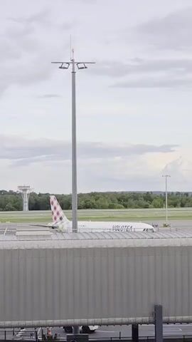 The video shows a Volotea airplane parked at an airport gate. The plane is white with red and black checkered tail markings, and the airline name "VOLOTEA" is visible on the side. In the background, a control tower and trees can be seen under a cloudy sky. In the foreground, part of the airport terminal is visible. The scene is filmed from a slightly elevated perspective, looking out towards the airplane.