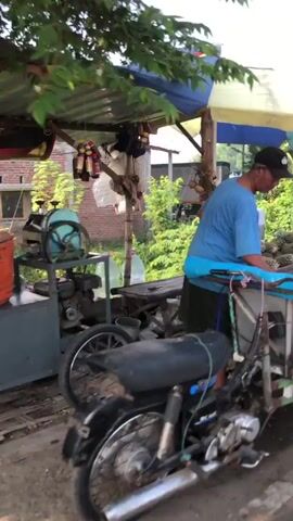 A person wearing a blue t-shirt and a black baseball cap is standing behind a stall. They are holding a pineapple in their hands and appear to be preparing it. The stall is laden with pineapples. In the background, there is a brick building and lush green vegetation. The scene suggests an outdoor market or roadside fruit stand.